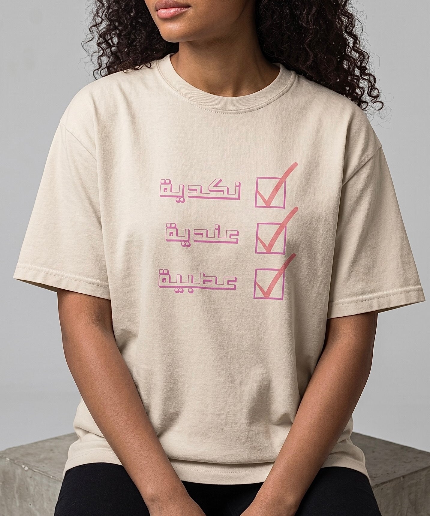 Model wearing a beige cotton T shirt with a pink Arabic checklist design featuring three lines of lettering from KIRO Clothing