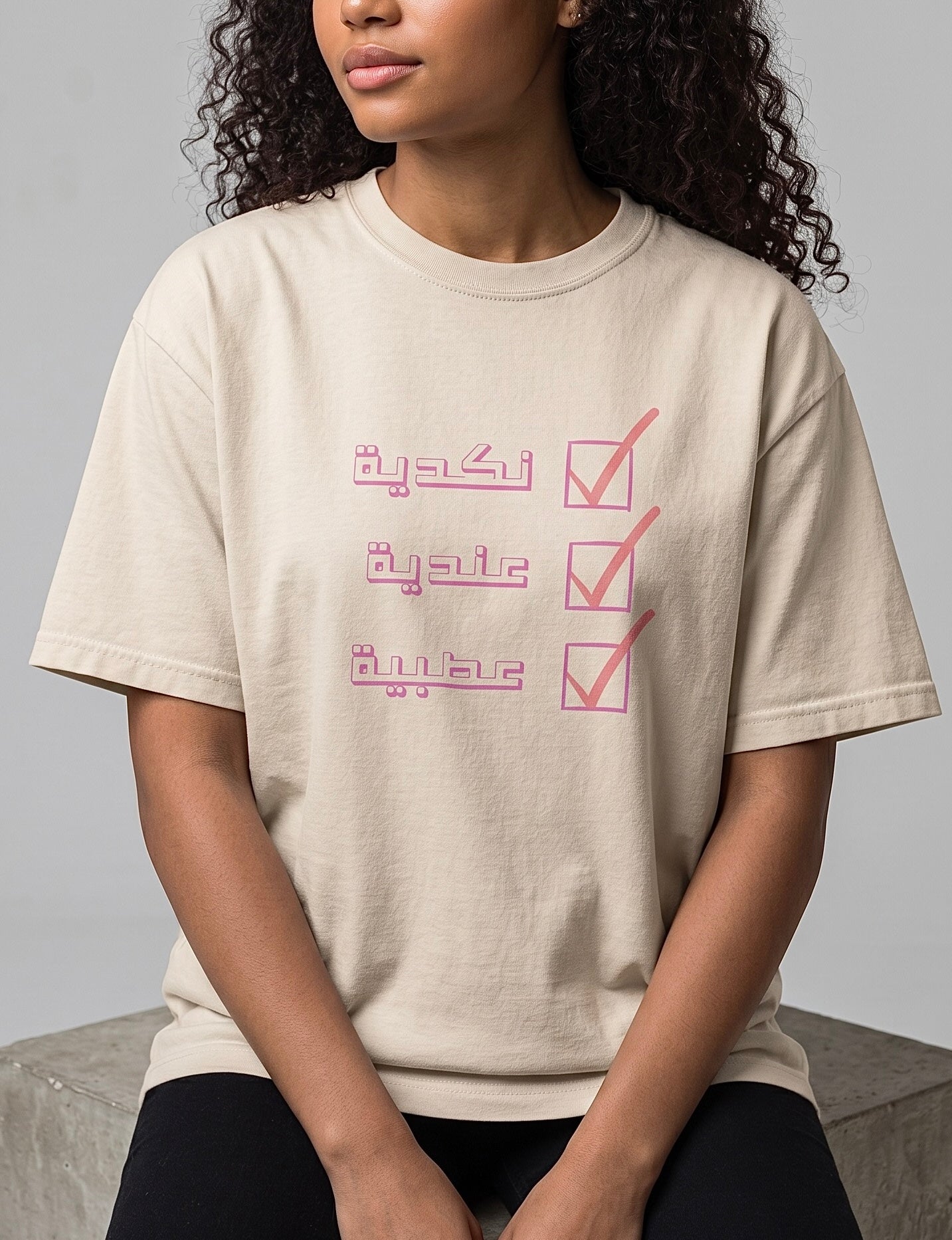 Model wearing a beige cotton T shirt with a pink Arabic checklist design featuring three lines of lettering from KIRO Clothing