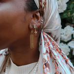Model wearing KIRO 18K gold plated stainless steel drop earrings featuring engraved Egyptian hieroglyph cartouche pendants. Also wearing crystal star earrings and a pearl necklace