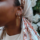 Model wearing KIRO 18K gold plated stainless steel drop earrings featuring engraved Egyptian hieroglyph cartouche pendants. Also wearing crystal star earrings and a pearl necklace