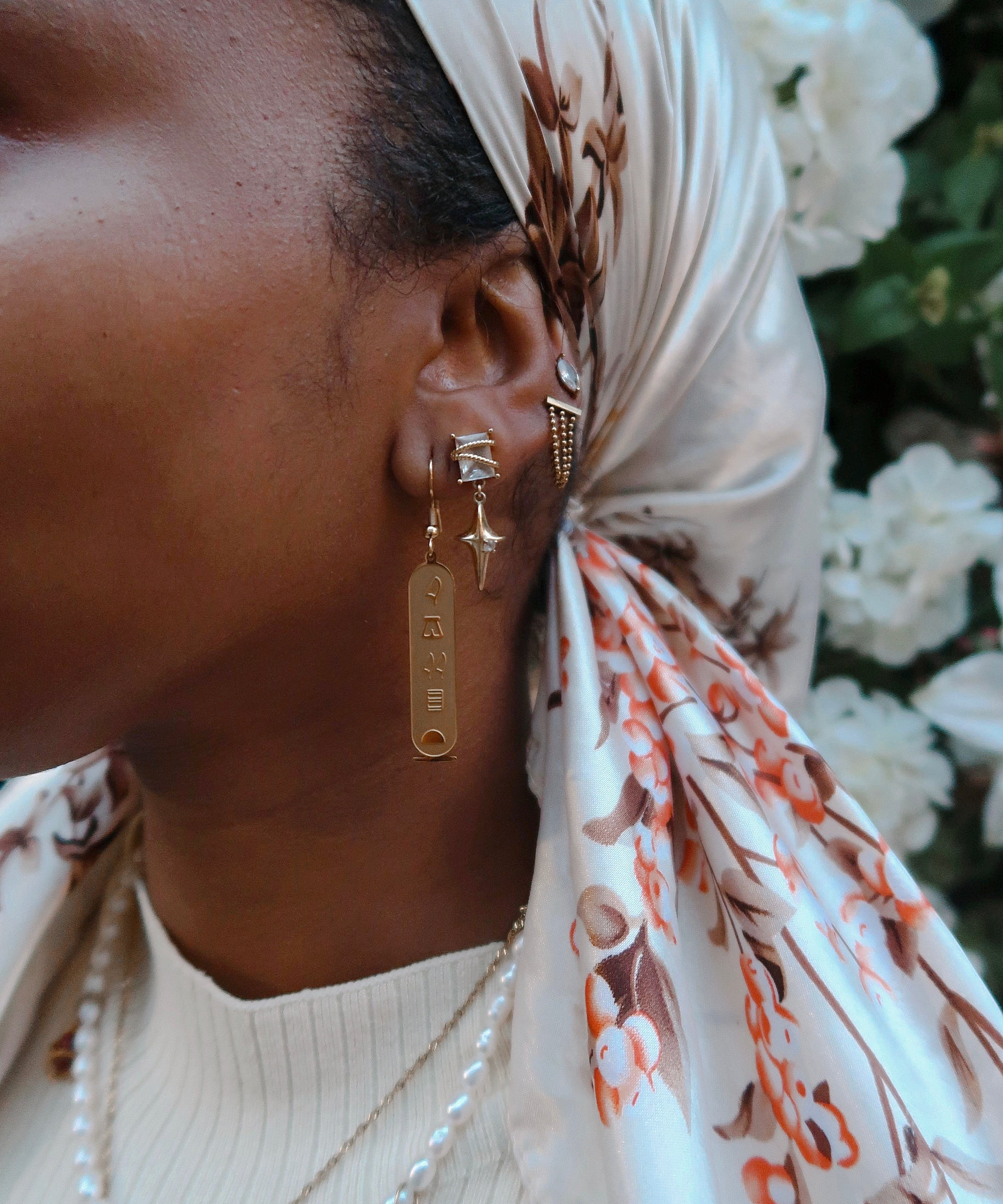 Model wearing KIRO 18K gold plated stainless steel drop earrings featuring engraved Egyptian hieroglyph cartouche pendants. Also wearing crystal star earrings and a pearl necklace